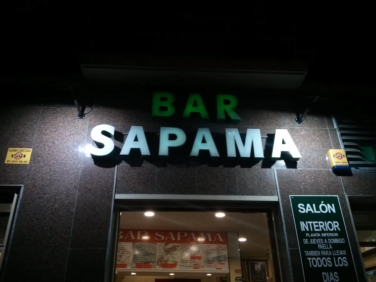 El rótulo de Bar Sapama encendido de noche: BAR en verde y SAPAMA en blanco sobre la fachada de granito, con el cartel del salón interior junto a la puerta